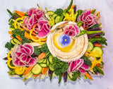 A beautifully arranged vegetable platter featuring a central bowl of creamy hummus topped with a yellow sauce and a flower garnish, surrounded by carved radishes, broccoli, cucumber slices, carrots, and yellow bell peppers on a white background.