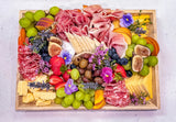 A large wooden charcuterie board filled with sliced cheeses, cured meats, grapes, figs, olives, strawberries, citrus slices, and small purple flowers, photographed from above on a light surface.