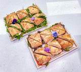 Two wooden trays holding multiple triangular sandwich cuts arranged on a light gray surface. The sandwiches are toasted bread with greens, fillings, and edible flower garnishes.