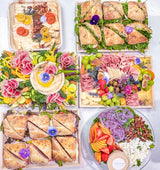 An overhead flat-lay of six large catering platters filled with gourmet food, including charcuterie boards, sandwiches, fruit, vegetables, cheese, crackers, and a small cup of dip on a white surface.