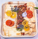 A wooden serving board filled with assorted crackers, sliced almonds, dried apricots, dried orange slices, lavender sprigs, and a small purple flower on a light surface.