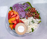 A clear glass bowl filled with a colorful fruit and cheese salad, featuring sliced strawberries, peaches, red onion, pecans, and crumbled feta cheese, with a small round container labeled 'Affinage artisanal spreads' placed in front. The bowl is photographed from above on a light gray marble surface.